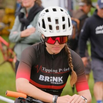 A female cyclist wearing a white helmet, red sunglasses, and a dark cycling jersey with "Tipping Point" logo. She's focused, with braided hair, in a lively outdoor setting.