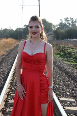 A woman in a vibrant red dress stands on railroad tracks, surrounded by nature. She has a confident smile, conveying elegance and confidence.