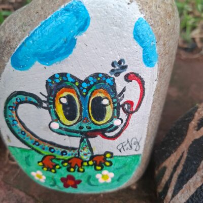 A colorful, cartoon-style lizard with large, expressive eyes is painted on a rock. The background features a blue sky and green grass with white flowers. The artwork conveys a playful and whimsical mood.