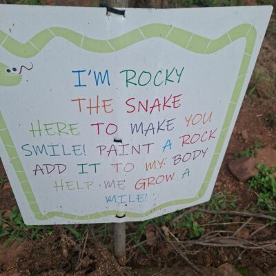 Sign with colorful text near rocky soil. It reads, "I'm Rocky the Snake. Here to make you smile! Paint a rock, add it to my body, help me grow a mile!"