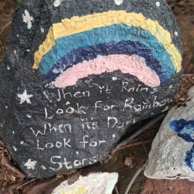 A painted rock features a colorful rainbow with stars. It displays a motivational message: "When it rains, look for rainbows; when it’s dark, look for stars."