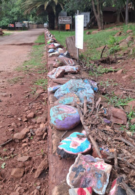 Colorful painted rocks line a dirt path, surrounded by scattered branches and greenery. Background signs suggest a rustic, natural setting.