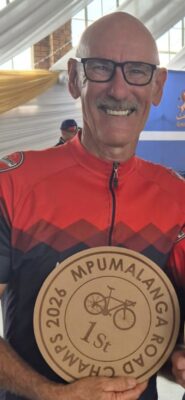 A smiling man in a red cycling jersey holds a "1st" place wooden plaque for the Mpumalanga 2026 Road Champs. The setting suggests a jubilant atmosphere.