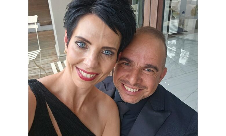 A smiling couple takes a selfie. The woman has short black hair, wearing a black dress; the man has short hair, wearing a dark jacket. They look happy.