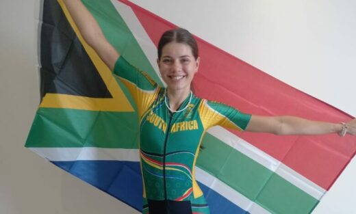 A woman wearing a green and yellow cycling jersey with "South Africa" printed on it smiles, arms outstretched, holding the South African flag behind her.