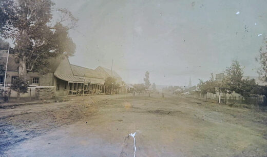 Vintage photograph of a dirt road flanked by rustic buildings and trees, evoking a nostalgic, serene atmosphere from a bygone rural era.