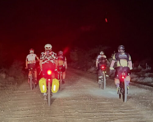 Five cyclists ride on a dirt road at night, illuminated by bike lights. The scene feels adventurous and communal, set against a dark backdrop.