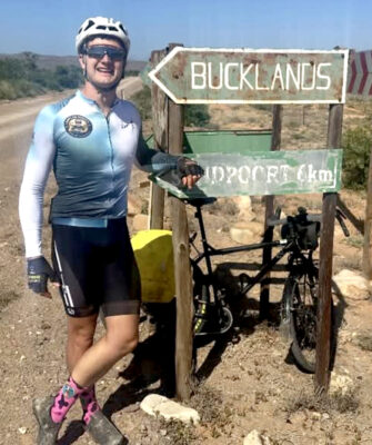 Cyclist in a blue jersey stands smiling beside a weathered road sign for "BucklandS," with a dusty bike in the background. Arid, sunny landscape.