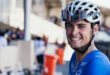 Cyclist in blue and yellow attire smiles beside a bike, holding handlebars. Background shows a race track with blurred spectators and bikes. Energetic mood.