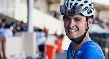 Cyclist in blue and yellow attire smiles beside a bike, holding handlebars. Background shows a race track with blurred spectators and bikes. Energetic mood.