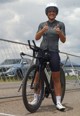 Cyclist in gear and helmet smiles, giving double thumbs-up beside a racing bike. Overcast sky and parked cars in the background convey a cheerful, competitive tone.