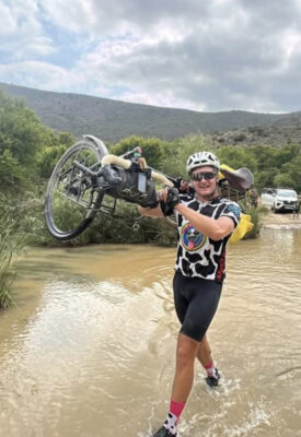 Smiling cyclist in cow-print jersey, holding bike above head while wading through muddy water. Green hills and cloudy sky in background convey adventure.