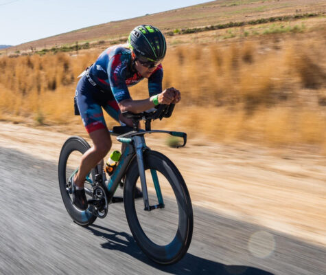 A cyclist in aerodynamic gear rides a time-trial bike on a rural road, with dry grass in the background. The scene exudes speed and focus.