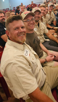 A group of men smiling and seated in a row, wearing casual outfits with name tags. The atmosphere is friendly and relaxed, suggesting a social or team event.
