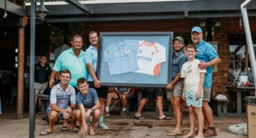 A group of smiling people gather outdoors on a wooden deck, holding a framed display featuring two sports jerseys. The mood is celebratory and casual.
