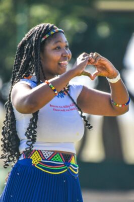 A woman smiling and forming a heart shape with her hands, dressed in colorful traditional African attire with beaded accessories, standing outdoors.