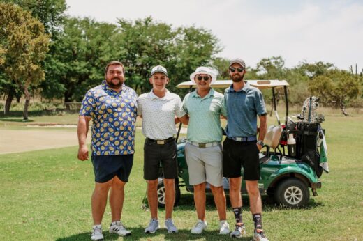 Four men stand on a golf course, smiling. They're casually dressed in colorful golf attire. A golf cart is behind them, with a lush green background.