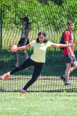 A woman in a yellow shirt and black leggings is captured mid-throw in a discus event. She stands on a grass field, with a net and spectators in the background.