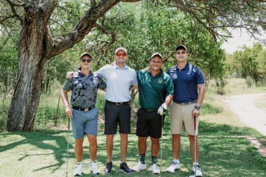 Four men in golf attire stand arm in arm on a lush golf course. They smile brightly under a large tree, surrounded by greenery. A path is visible on the right.