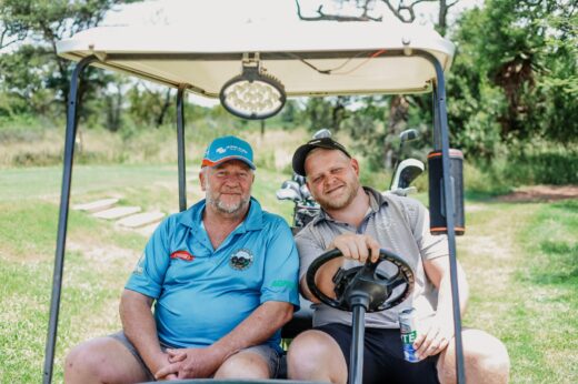 Two men are sitting in a golf cart on a sunny day, smiling. The older man wears a blue shirt and cap, the younger man a striped shirt and cap. Lush greenery surrounds them.