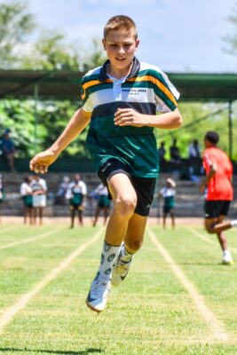 A young boy is captured mid-jump in a race on a grassy sports field, wearing a green-striped uniform and patterned socks, with determination on his face.