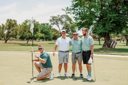 Four men on a golf course pose happily. One kneels by the flag, while the others stand close together. The day is sunny with lush green trees.