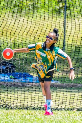A young athlete enthusiastically throws a discus, standing on grass with a net backdrop. Wearing a colorful outfit and sunglasses, she exudes joy and energy.