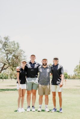 Four individuals stand together on a golf course, smiling. They are dressed in casual golf attire. The background features trees and a cloudy sky.