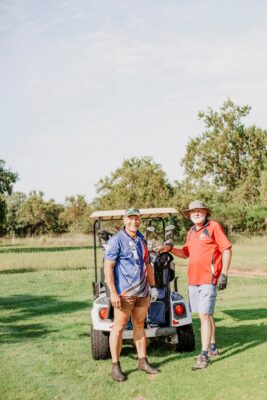 Two men stand smiling in front of a golf cart on a sunny golf course. One wears a blue shirt; the other a red one. Green trees surround them under a clear sky.