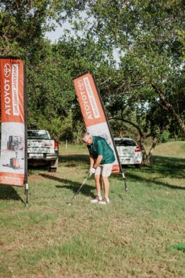 A golfer prepares to swing on a grassy field flanked by Toyota banners, with parked vehicles and trees in the background, under a sunny sky.