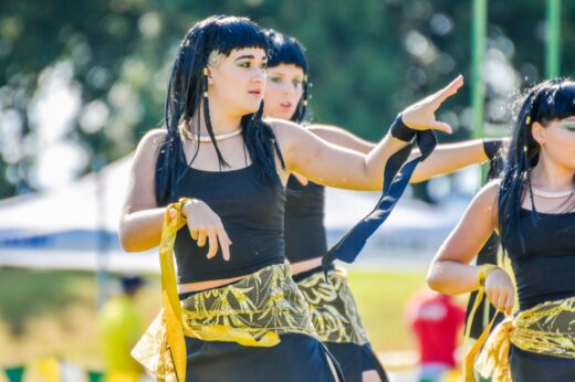 Performers wearing black outfits and golden sashes dance energetically outdoors. Their focused expressions convey concentration and excitement.
