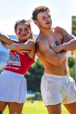 Two young people pose confidently outdoors. The woman wears a red USA tank top with face paint, while the man is shirtless, both smiling. Bright, sunny day.