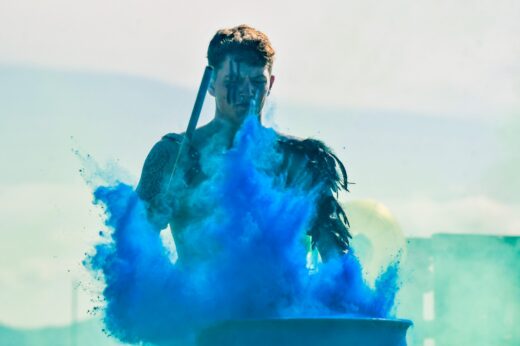 A person in tribal attire, holding a stick, is surrounded by a burst of vibrant blue powder, set against a bright, hazy background. The mood is intense.