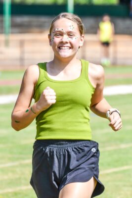 Smiling teenager with face paint runs on grass, wearing a green tank top and black shorts. Sunny day, background blurred, conveying joy and energy.