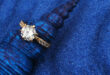 A sparkling diamond engagement ring with a gold band rests elegantly on a textured, deep blue shell, set against a vibrant blue sand background.