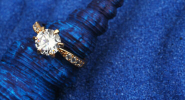 A sparkling diamond engagement ring with a gold band rests elegantly on a textured, deep blue shell, set against a vibrant blue sand background.