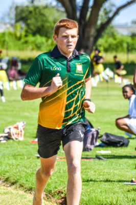 A young man runs on a grassy field, wearing a green and yellow sports jersey. Trees and other participants are blurred in the sunny background.