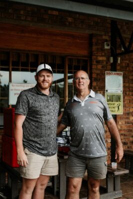 Two men stand smiling in front of a brick building. Both wear casual shorts and shirts. Behind them are caution signs and a window with grills.