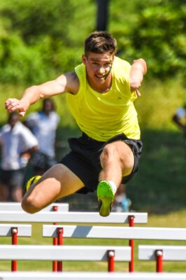 A male athlete in a yellow tank top leaps over a hurdle with determination, set against a blurred, sunny, outdoor background.