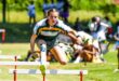 A young athlete leaps mid-air over a hurdle on a sunny track field, focused and determined. She wears a sports uniform with green and yellow stripes, with spectators blurred in the background.