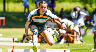 A young athlete leaps mid-air over a hurdle on a sunny track field, focused and determined. She wears a sports uniform with green and yellow stripes, with spectators blurred in the background.