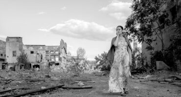 A woman in a long floral dress, with a contemplative expression, walks through an overgrown, crumbling urban landscape, conveying solitude and resilience.