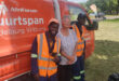 Two smiling workers in reflective vests and a woman stand in front of an orange truck with "AfriForum" text. Bright, sunny day, casual and cheerful mood.
