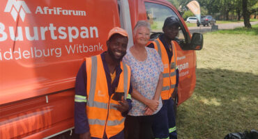 Two smiling workers in reflective vests and a woman stand in front of an orange truck with "AfriForum" text. Bright, sunny day, casual and cheerful mood.