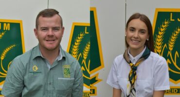 Two individuals smiling, each in front of a green and yellow emblem with wheat graphics. The man wears a green shirt; the woman, a white shirt with a scarf.