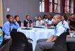 A group of students in school uniforms sit attentively around a round table with bottled water, in a room with a formal setting and focused atmosphere.