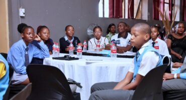A group of students in school uniforms sit attentively around a round table with bottled water, in a room with a formal setting and focused atmosphere.