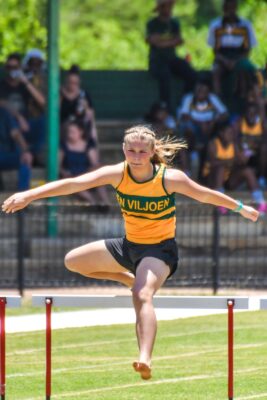 A young female athlete in a yellow and black uniform is mid-air, clearing a hurdle during a race. Spectators in the background watch attentively.