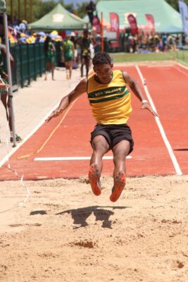 An athlete in a yellow and green uniform mid-air in a long jump, focused and determined. A red track and onlookers in the sunny background.
