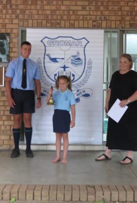 A young girl stands holding a bell between a smiling student and an adult on a stage with a "Gromar" school banner, conveying pride and accomplishment.